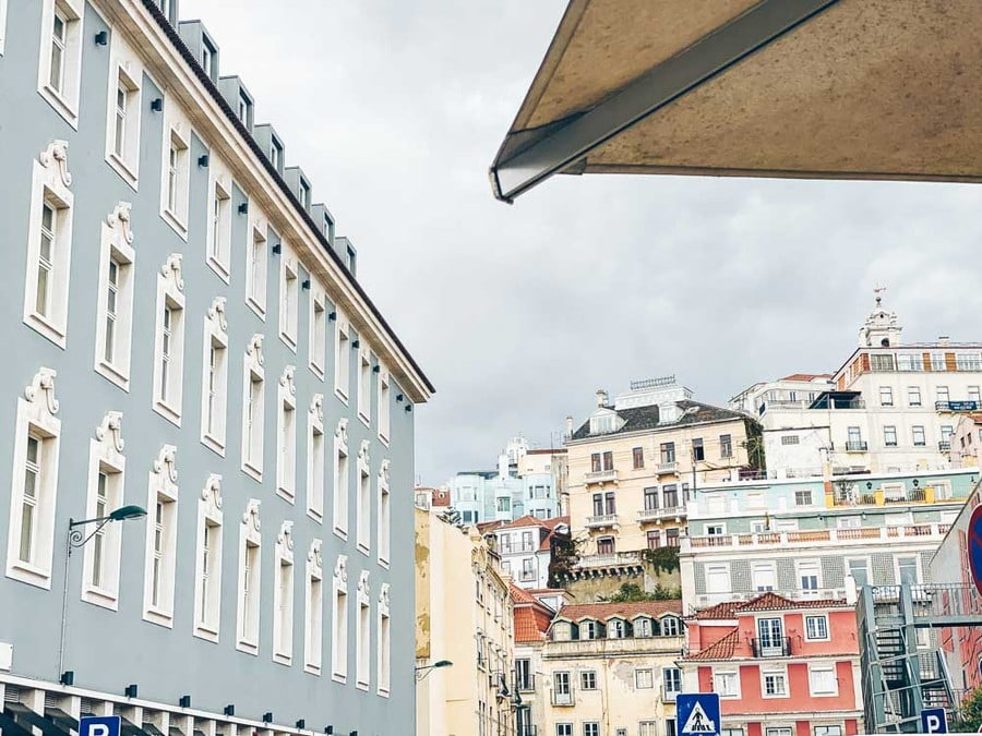 A pale blue building with ornate window frames and a hillside of colourful houses behind it in Lisbon