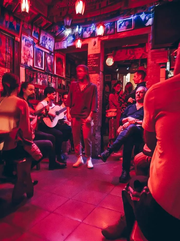 A guitarist and singer performing fado among a crowd in a small red-lit tasca in Lisbon