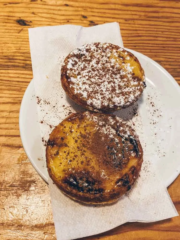 Two golden pastéis de nata with caramelised tops on a paper napkin on a wooden counter in Lisbon