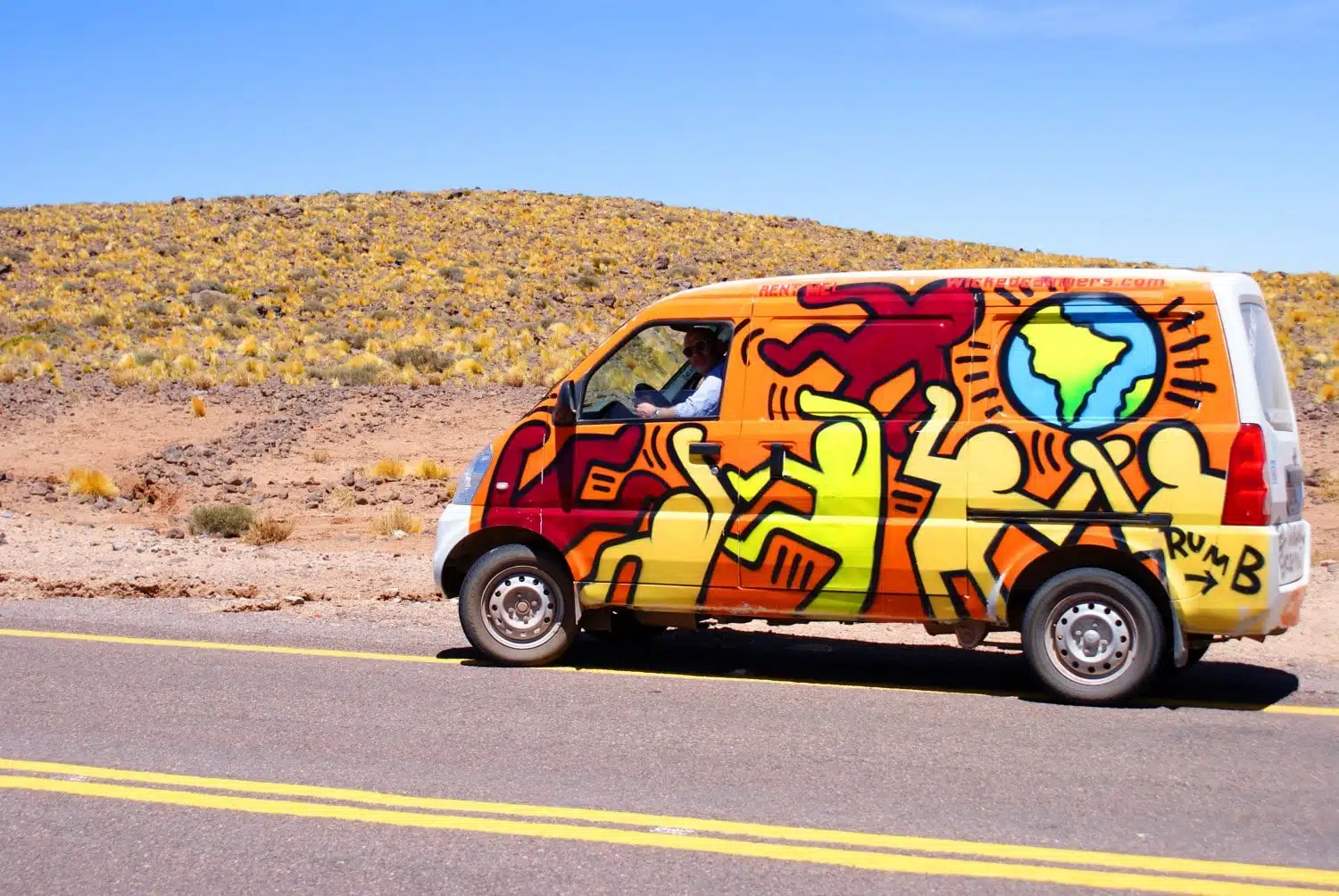 Brightly painted campervan covered in cartoon figures, parked on a paved desert road in the Atacama