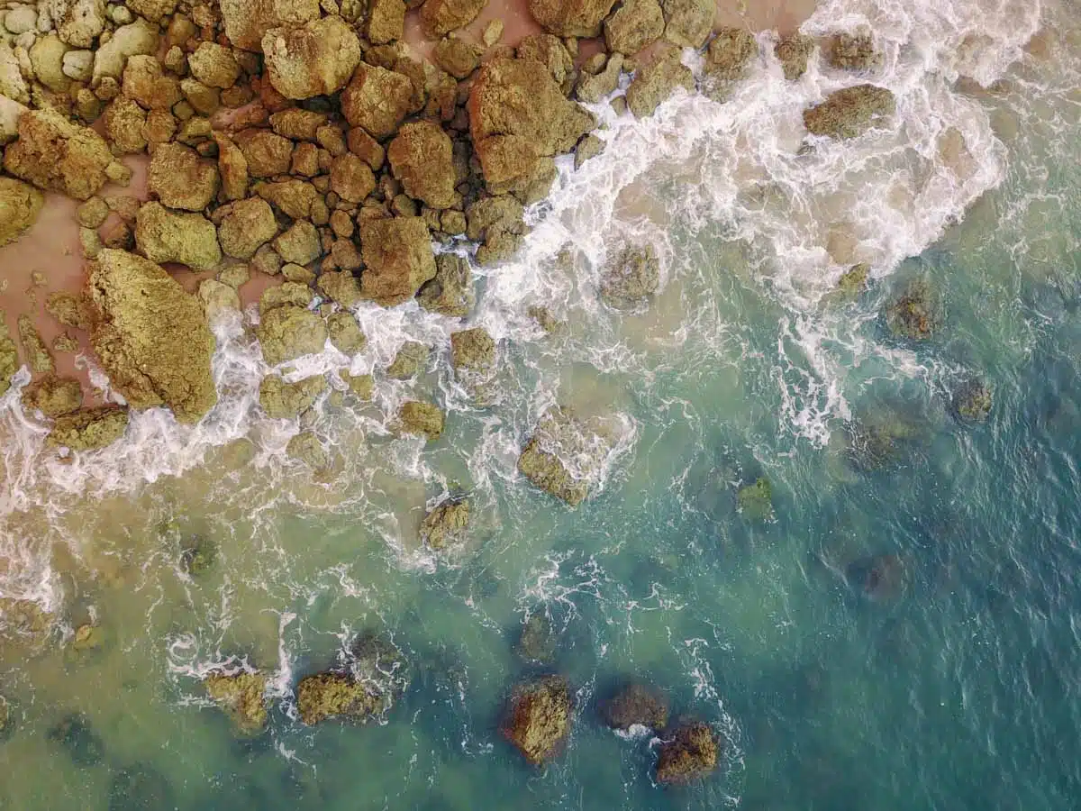 Drone view of white surf washing over golden rocks and clear green water at the foot of the cliffs on the Algarve coast