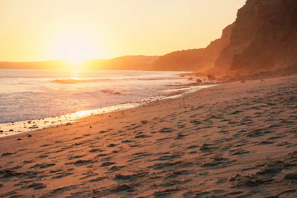 Low sun over a wide sandy beach with footprints in the sand and golden cliffs on the western Algarve coast