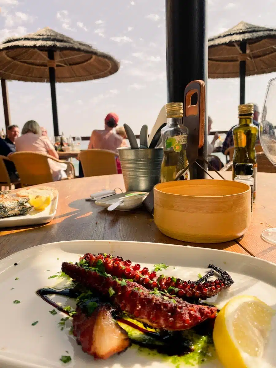 Plate of grilled octopus with lemon on a terrace table under straw parasols on the Algarve coast
