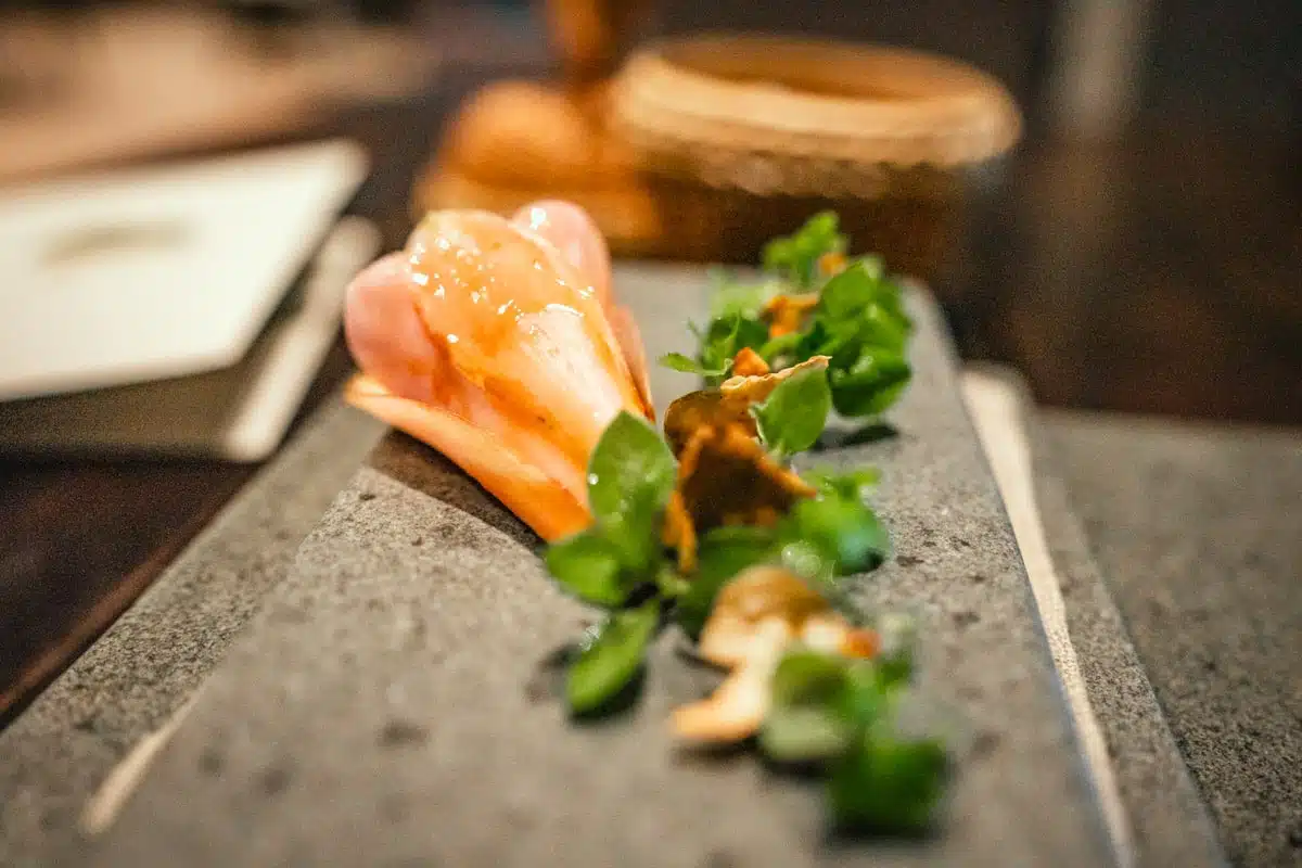 Pale pink petal-shaped bite with herbs and slivers of mushroom on a long stone slab at Boragó