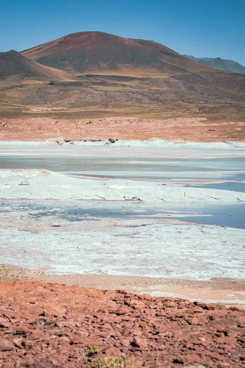 Brown volcano rising above white salt crust and turquoise water on the Salar de Talar, with red ground in front