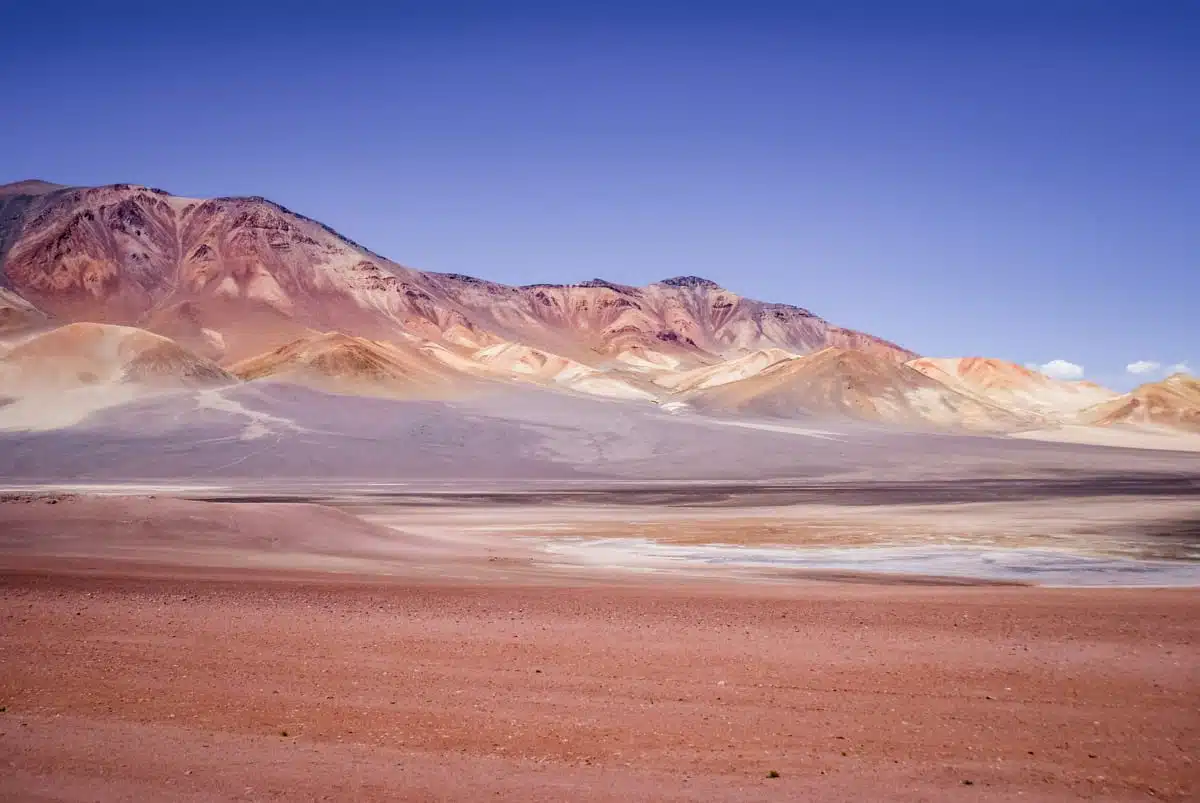 Hillside streaked with rust, cream and violet minerals above a red plain beside Ruta 27 in the Atacama