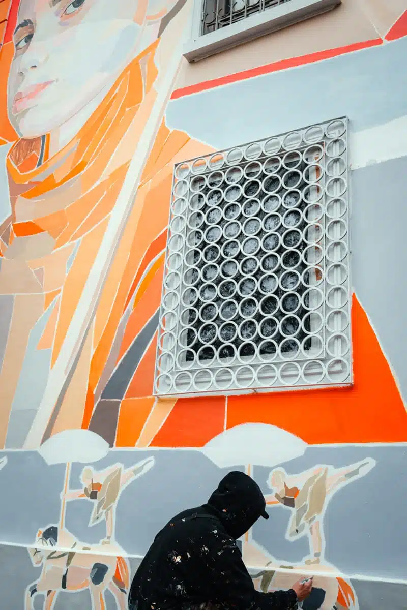 Artist painting a new mural of a woman in an orange scarf on a pastel house front in Bellavista, Santiago