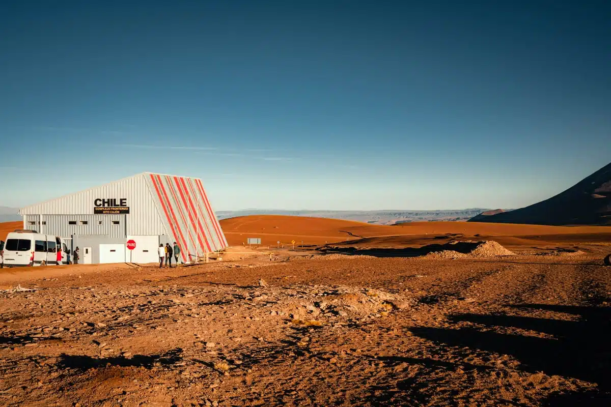 Striped Chilean border building and parked minibuses on the red-brown altiplano at the Bolivian frontier