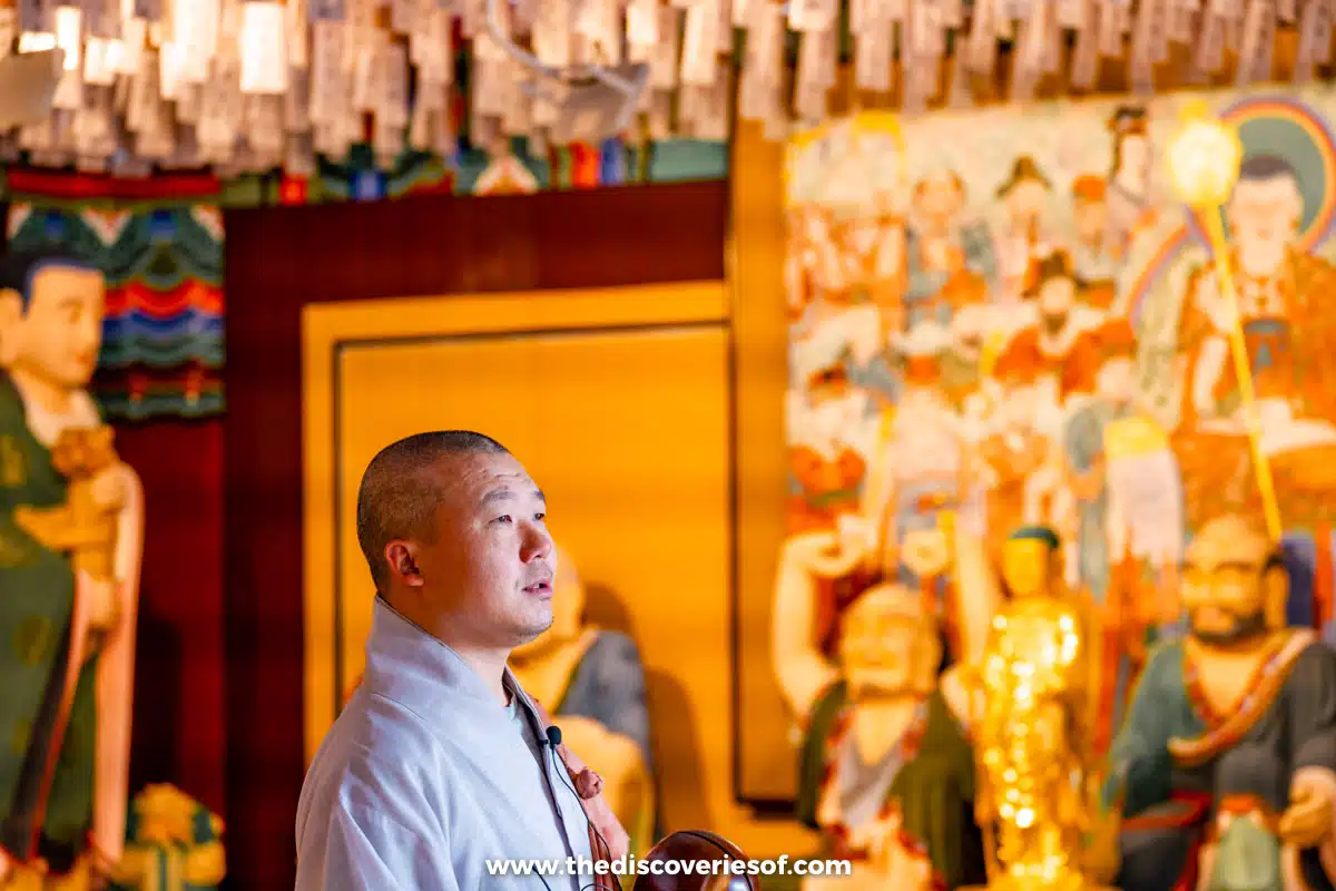 A monk in grey robes with a headset microphone in front of painted Buddhist statues and murals inside a hall at Haedong Yonggungsa