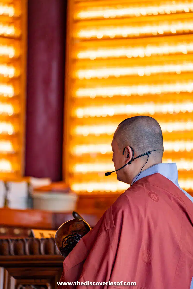 A monk in a red robe striking a wooden moktak in front of a wall of small glowing lights inside a hall at Haedong Yonggungsa