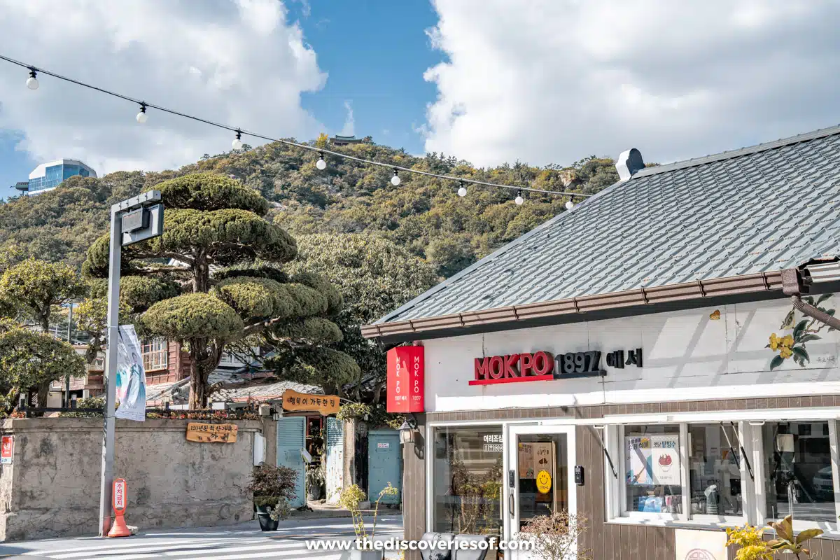 A white shop with a red Mokpo 1897 sign and a sculpted pine below wooded Yudalsan in the old town