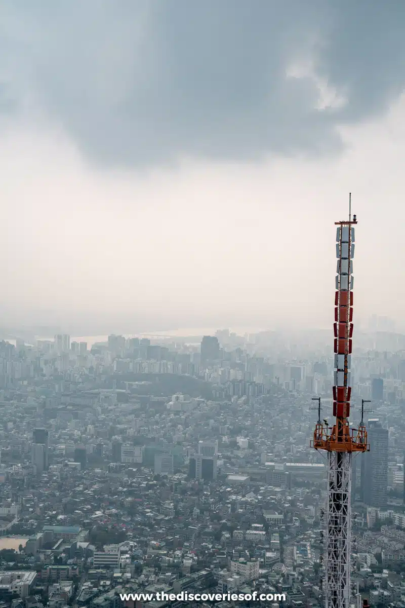 A red-and-white transmission mast standing over the hazy sprawl of Seoul, seen from a window at N Seoul Tower