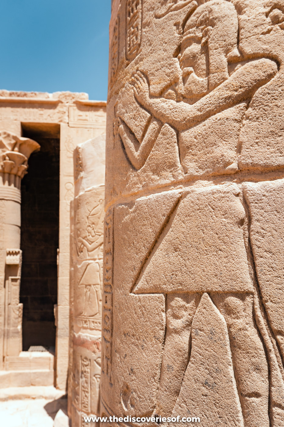 A sandstone column carved with a kilted figure raising its arms, beside a doorway at Philae Temple, Aswan