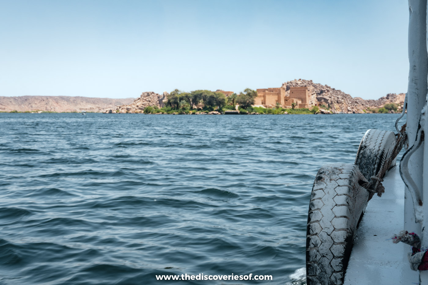 Philae Temple on its island seen across choppy blue water from the side of a motorboat near Aswan