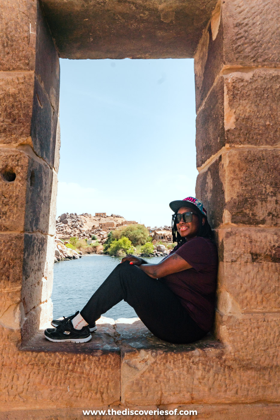 Julianna in trainers and trousers sitting in a stone window at Philae Temple, with the Nile behind her