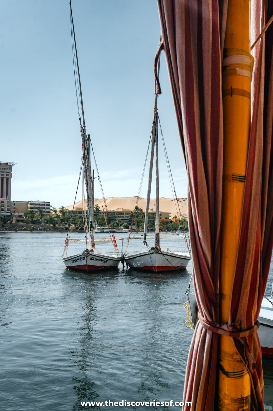 Two white-sailed feluccas moored on the Nile in Aswan, seen from under a striped awning