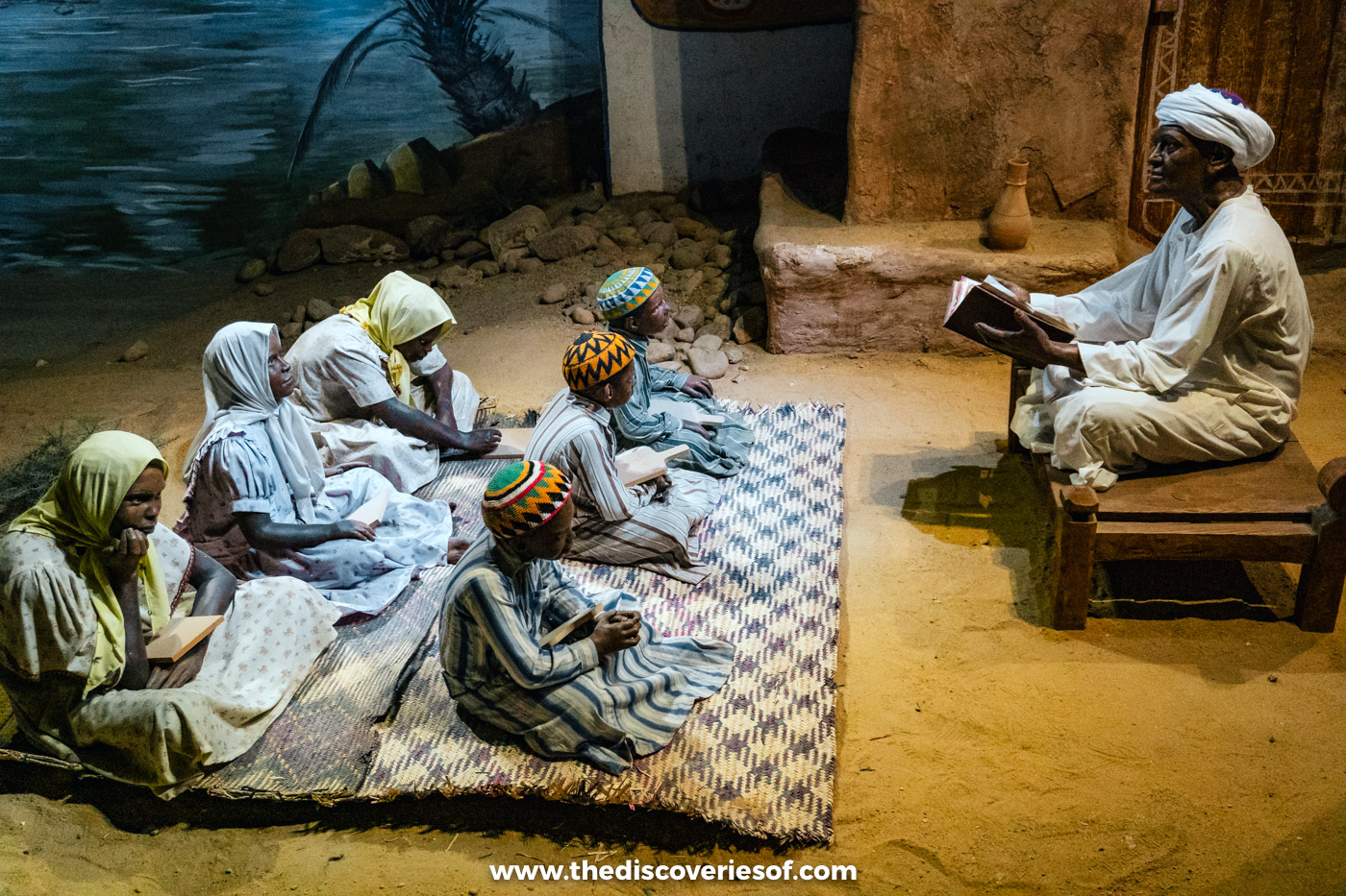 A life-size diorama of Nubian children sitting on mats in front of their teacher at a village Quran school