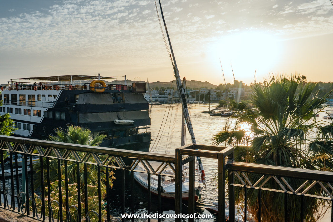 A multi-deck Nile cruise boat moored in Aswan, seen from a terrace with palms and a felucca mast