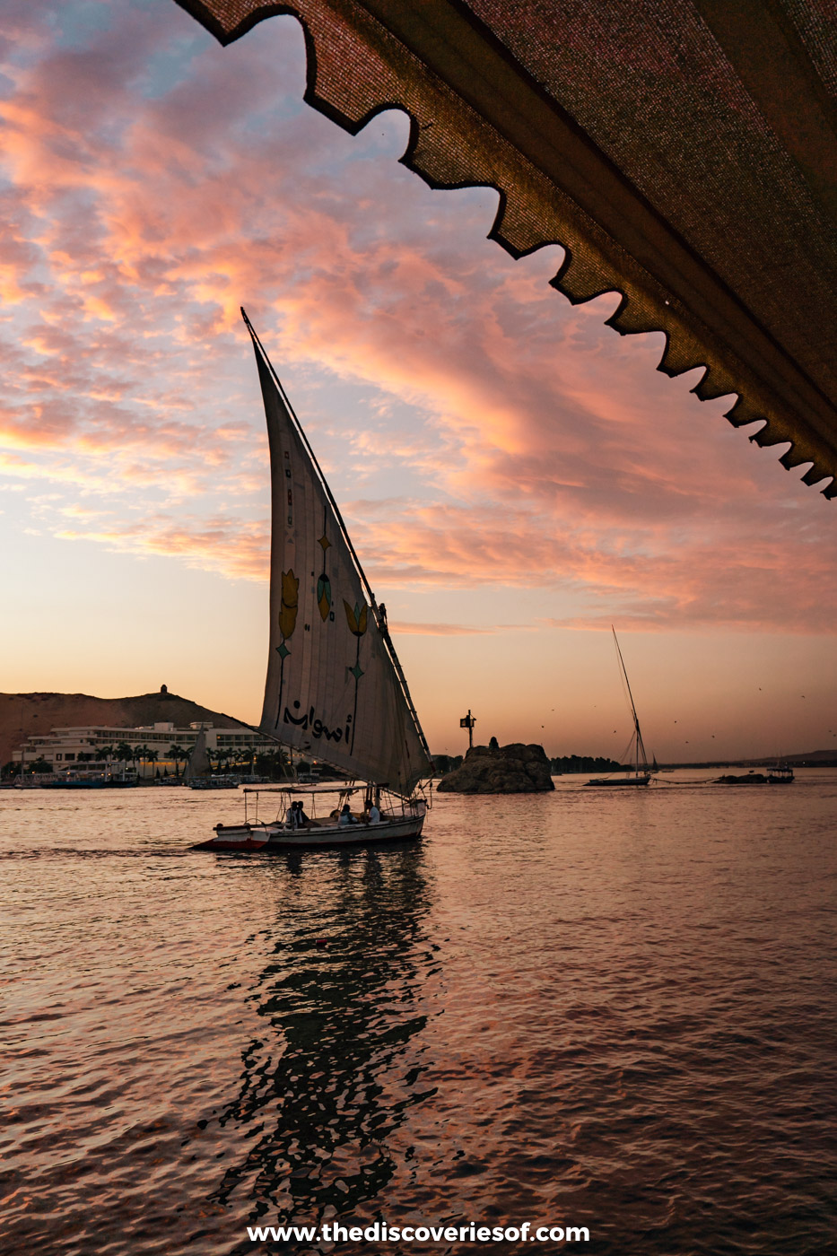 A felucca with a tall white sail gliding across the Nile under a pink sunset sky in Aswan