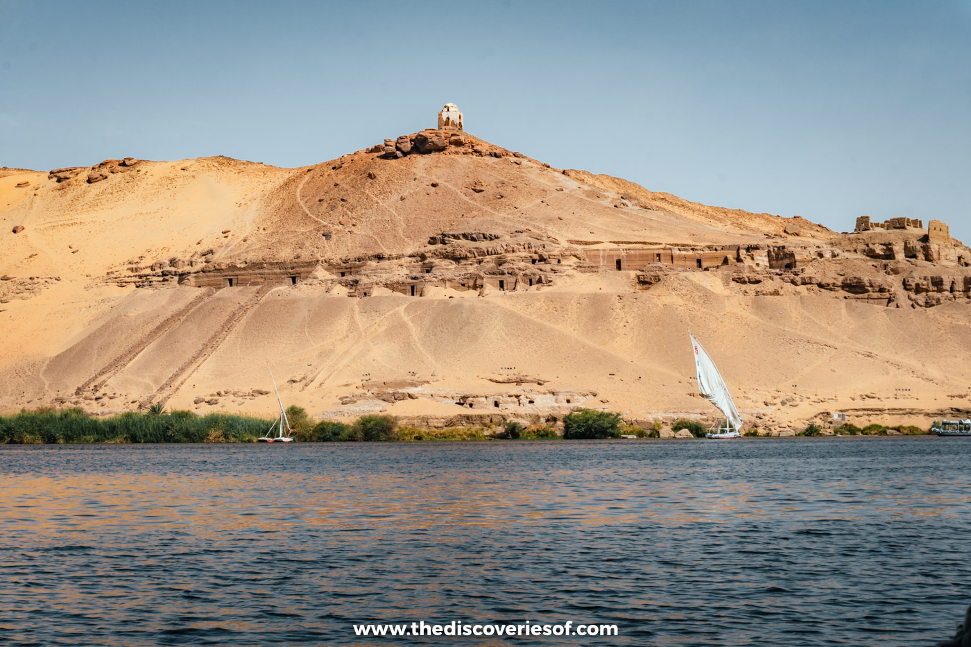 The Tombs of the Nobles cut into the sandy West Bank hill below the Qubbet el-Hawa dome, with a felucca