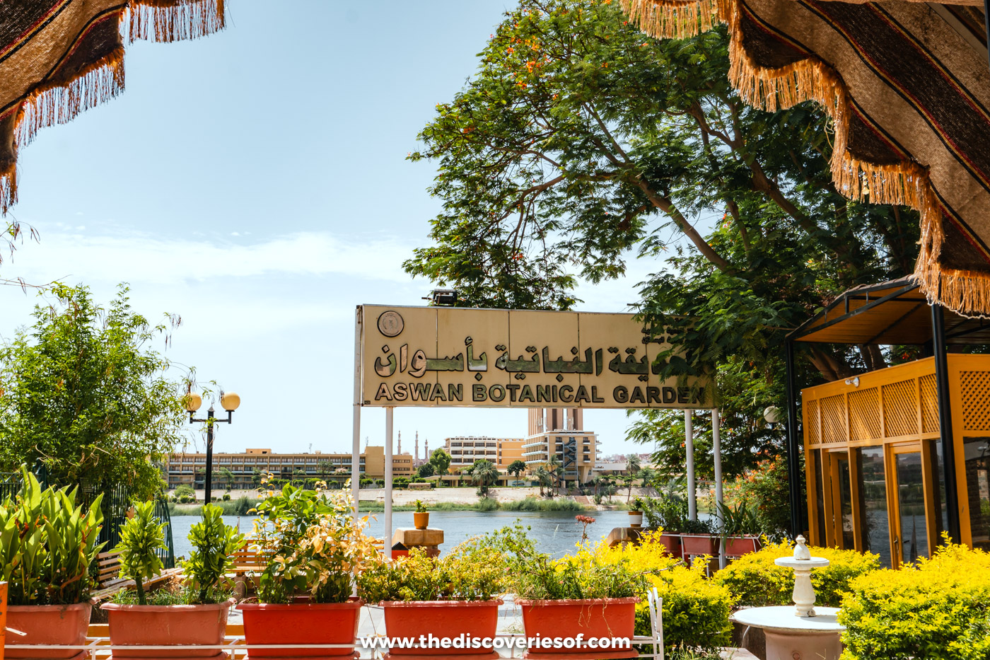 The Aswan Botanical Garden sign beside a small riverside café with potted flowers and the Nile behind