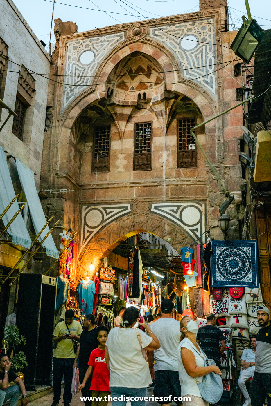 Shoppers passing beneath a tall carved stone Mamluk gateway hung with lanterns and textiles in Khan el-Khalili