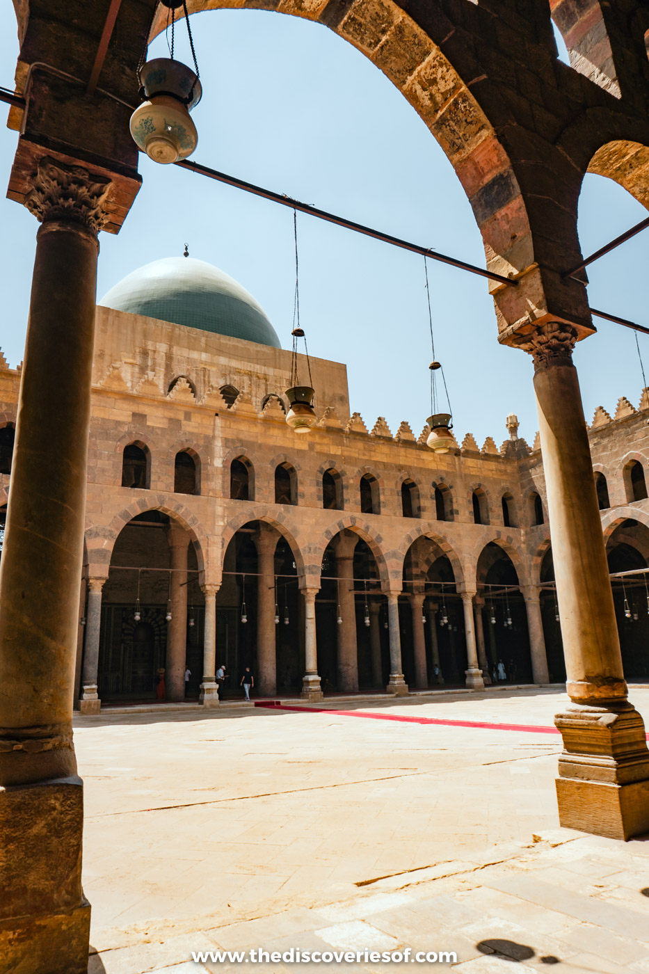 Striped stone arches and hanging lamps around the courtyard of the Mosque of al-Nasir Muhammad, with its green dome