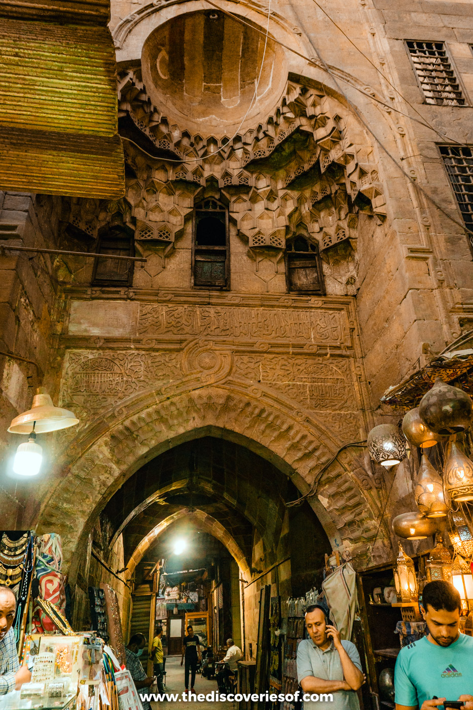 A carved Mamluk stone gateway above a lantern-lit lane of shops in the Khan el-Khalili bazaar, Cairo