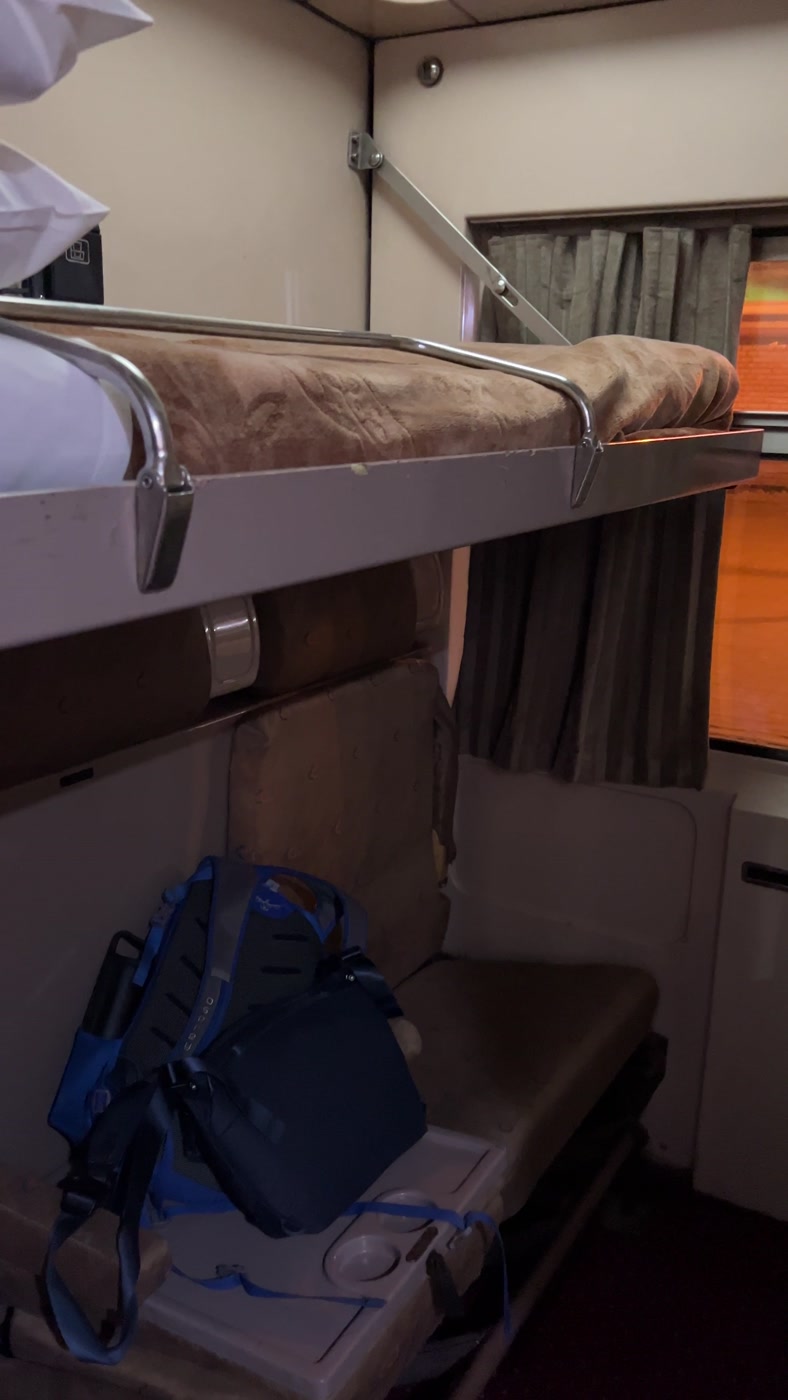 A two-berth sleeper train cabin with the upper bunk made up, seats below, a backpack and curtained window