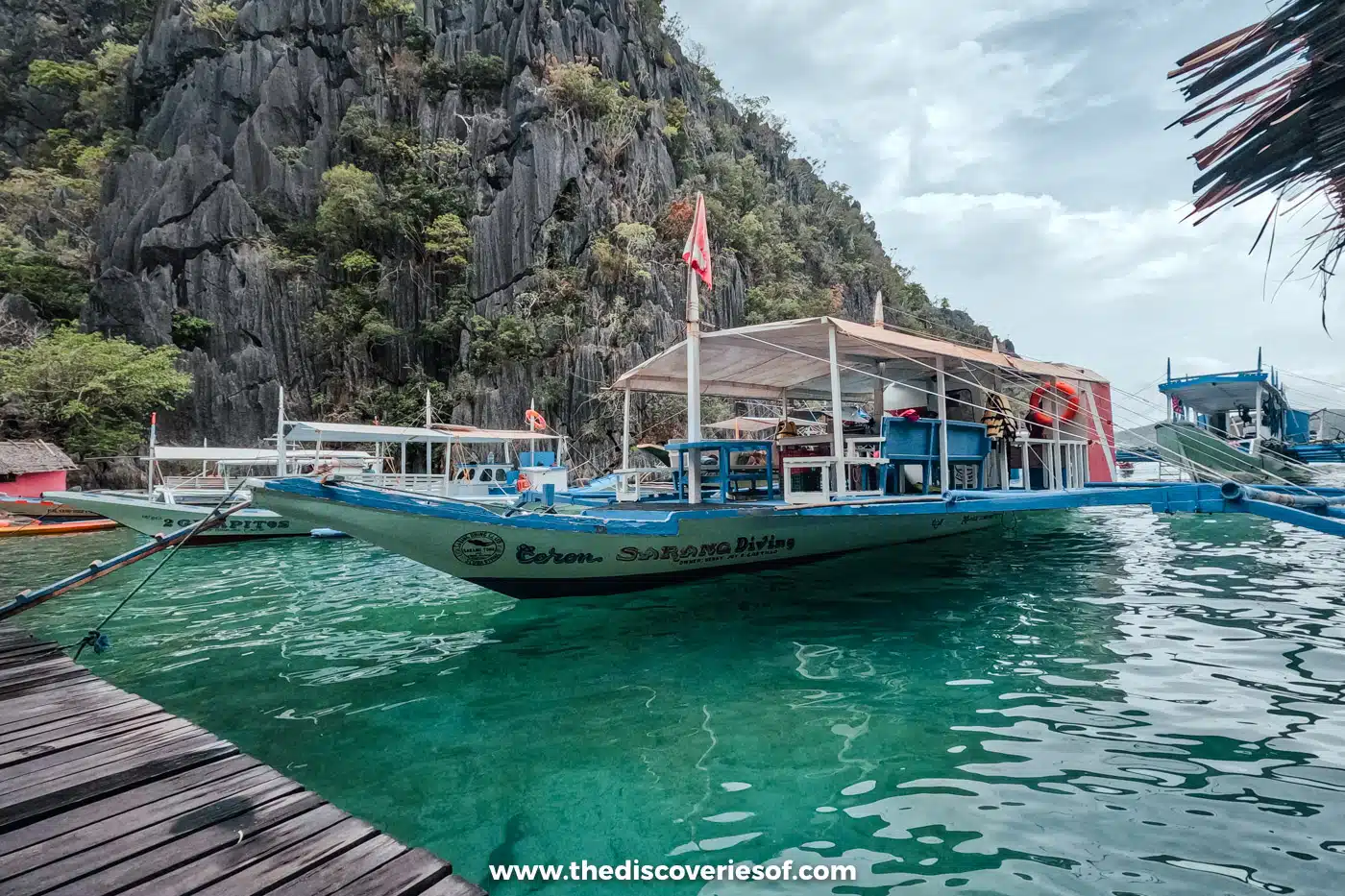 Bangka boat moored at a wooden jetty below a jagged limestone cliff on a Coron island-hopping tour