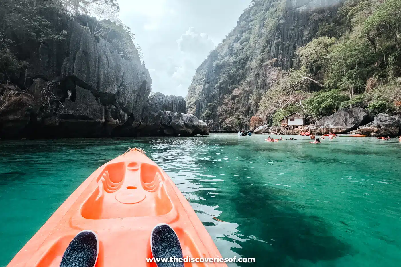 View from the bow of an orange kayak towards limestone cliffs and swimmers in the Twin Lagoon, Coron