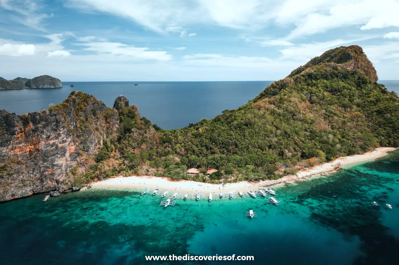 Aerial view of Helicopter Island's long white beach below a green ridge with boats in turquoise water, El Nido