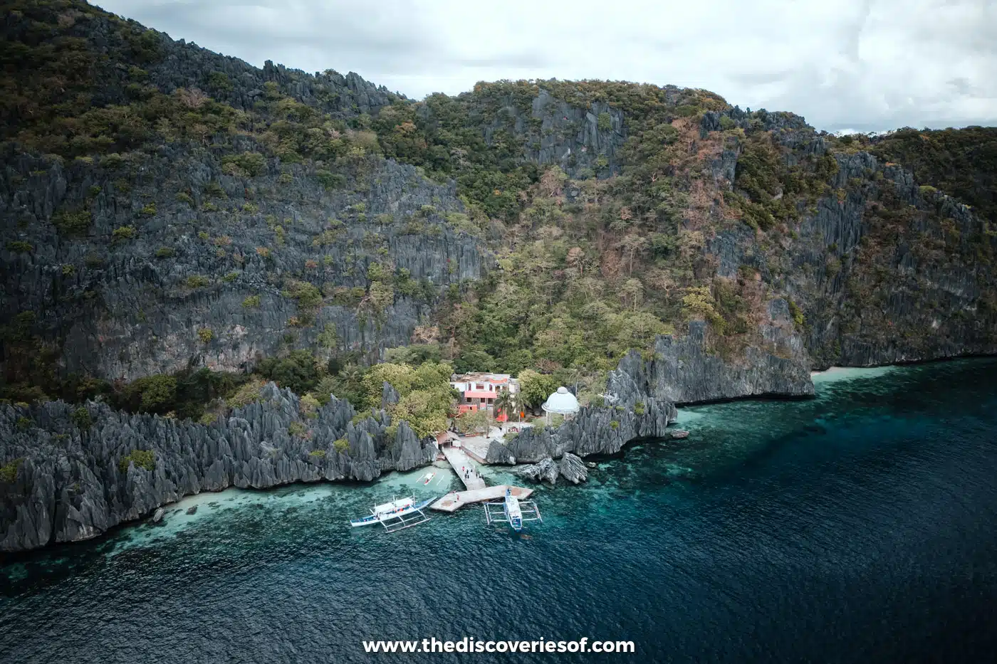 Aerial view of Matinloc Shrine's white buildings and jetty in a small cove below grey limestone cliffs, El Nido