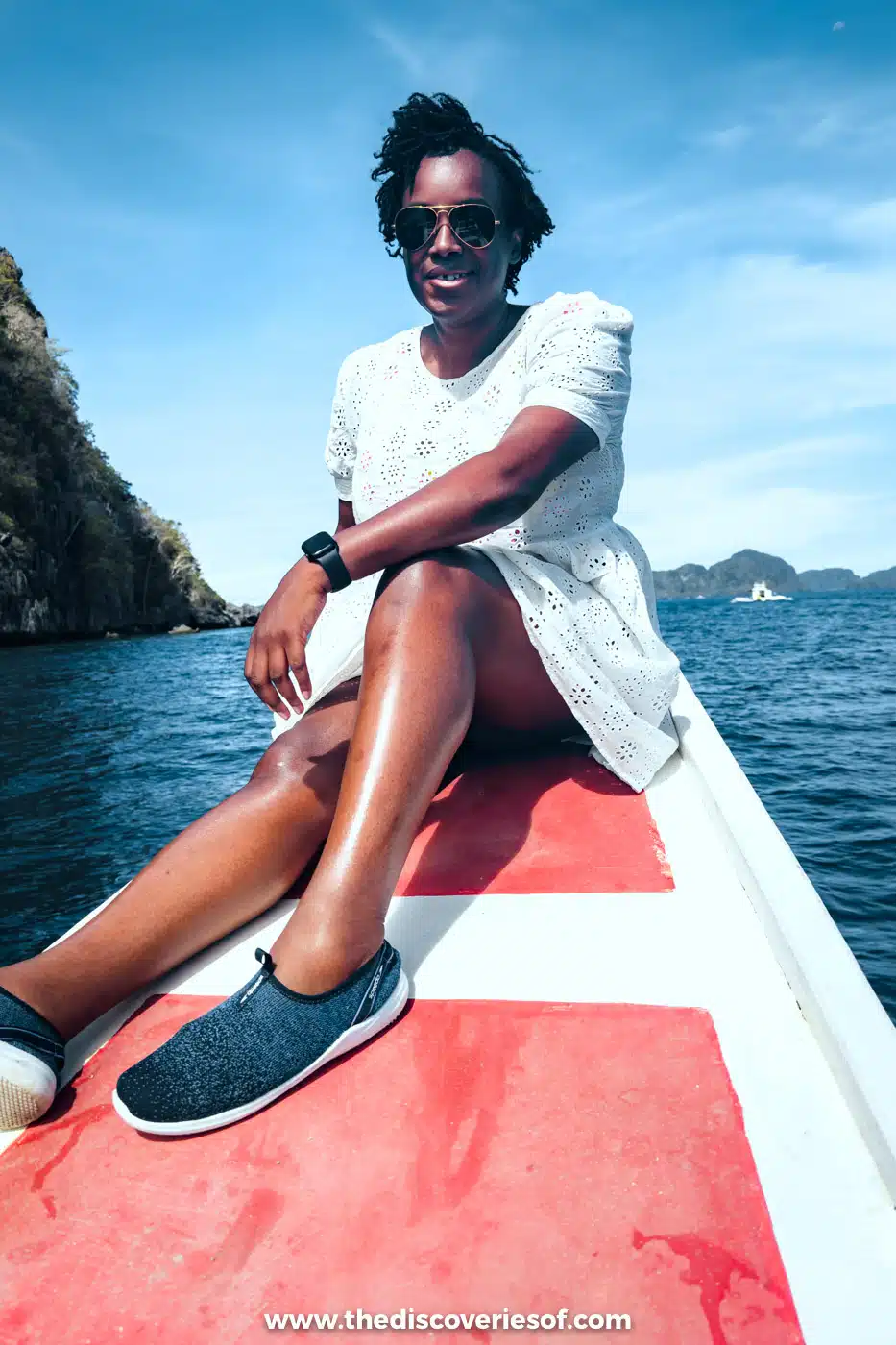 Julianna in a white dress sitting on the red and white bow of a bangka boat on the open sea near El Nido