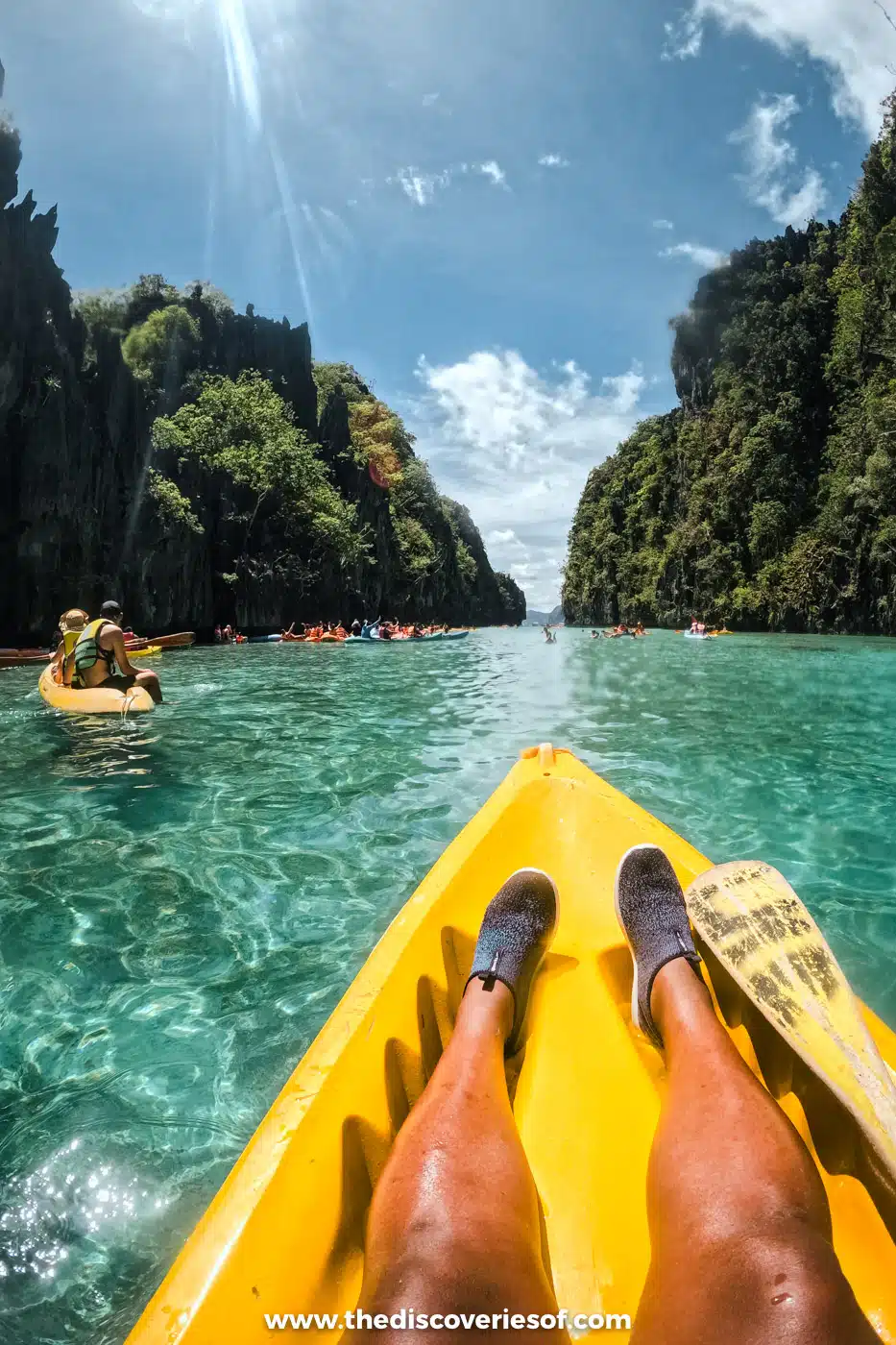 Julianna's legs in a yellow kayak gliding over clear turquoise water towards limestone cliffs in the Big Lagoon, El Nido