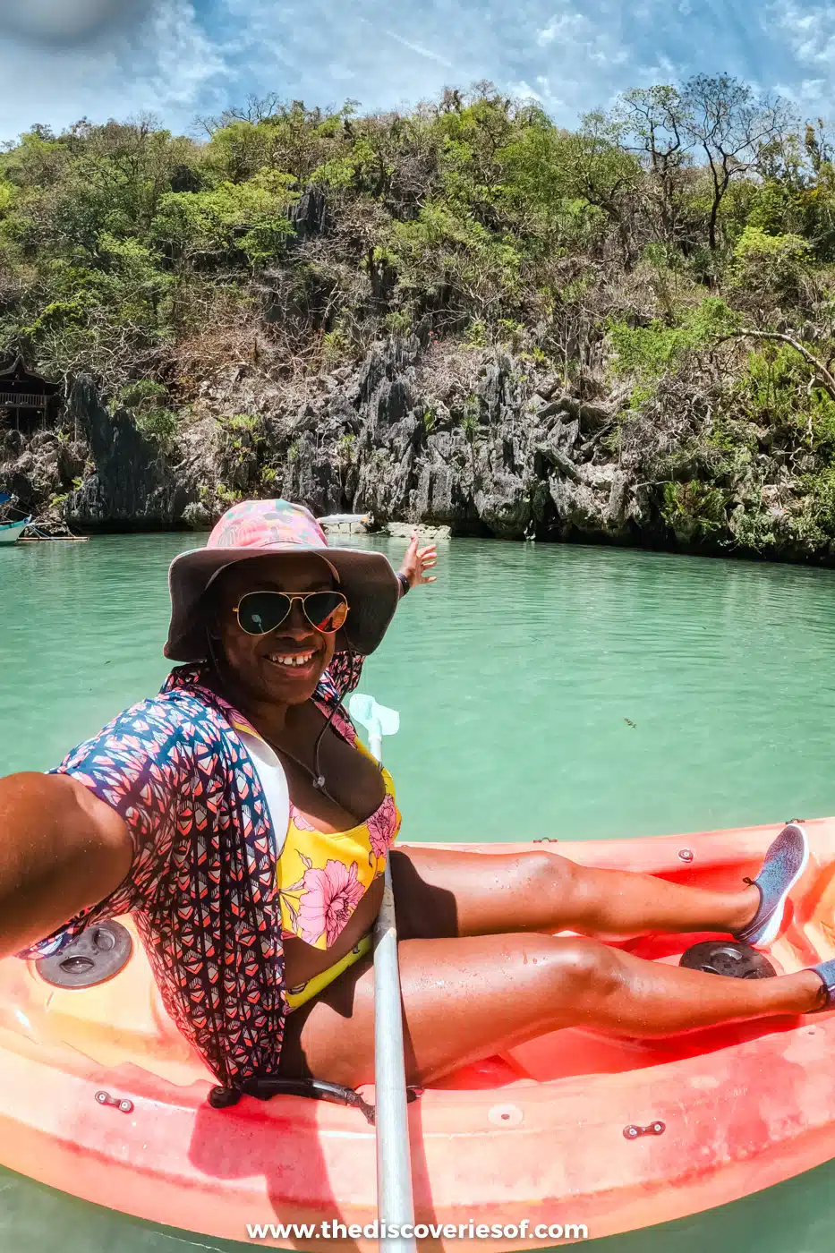 Julianna in a sun hat paddling a coral-coloured kayak in a green lagoon below limestone cliffs, El Nido