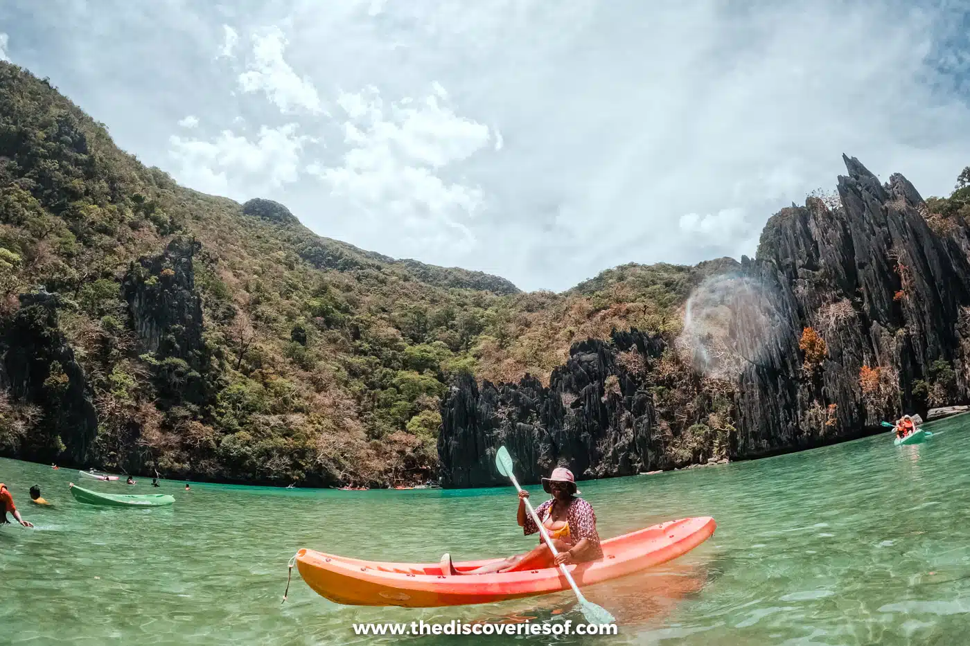 Kayaker in an orange kayak on the calm green water of Cadlao Lagoon below jagged cliffs, El Nido