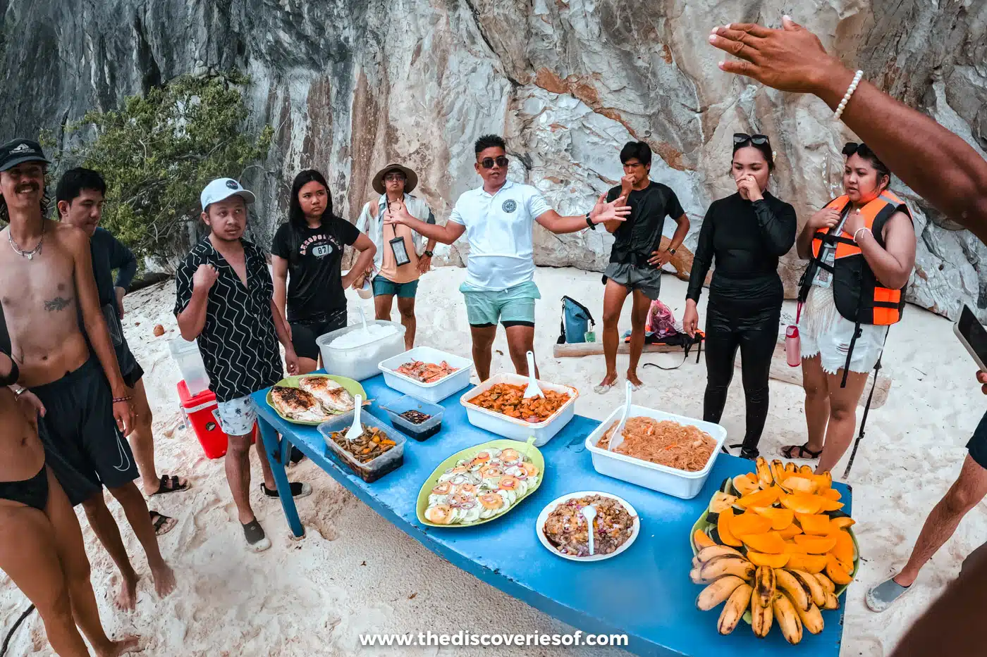 Island-hopping group gathered round a table of grilled fish, rice and fruit on a white-sand beach, El Nido