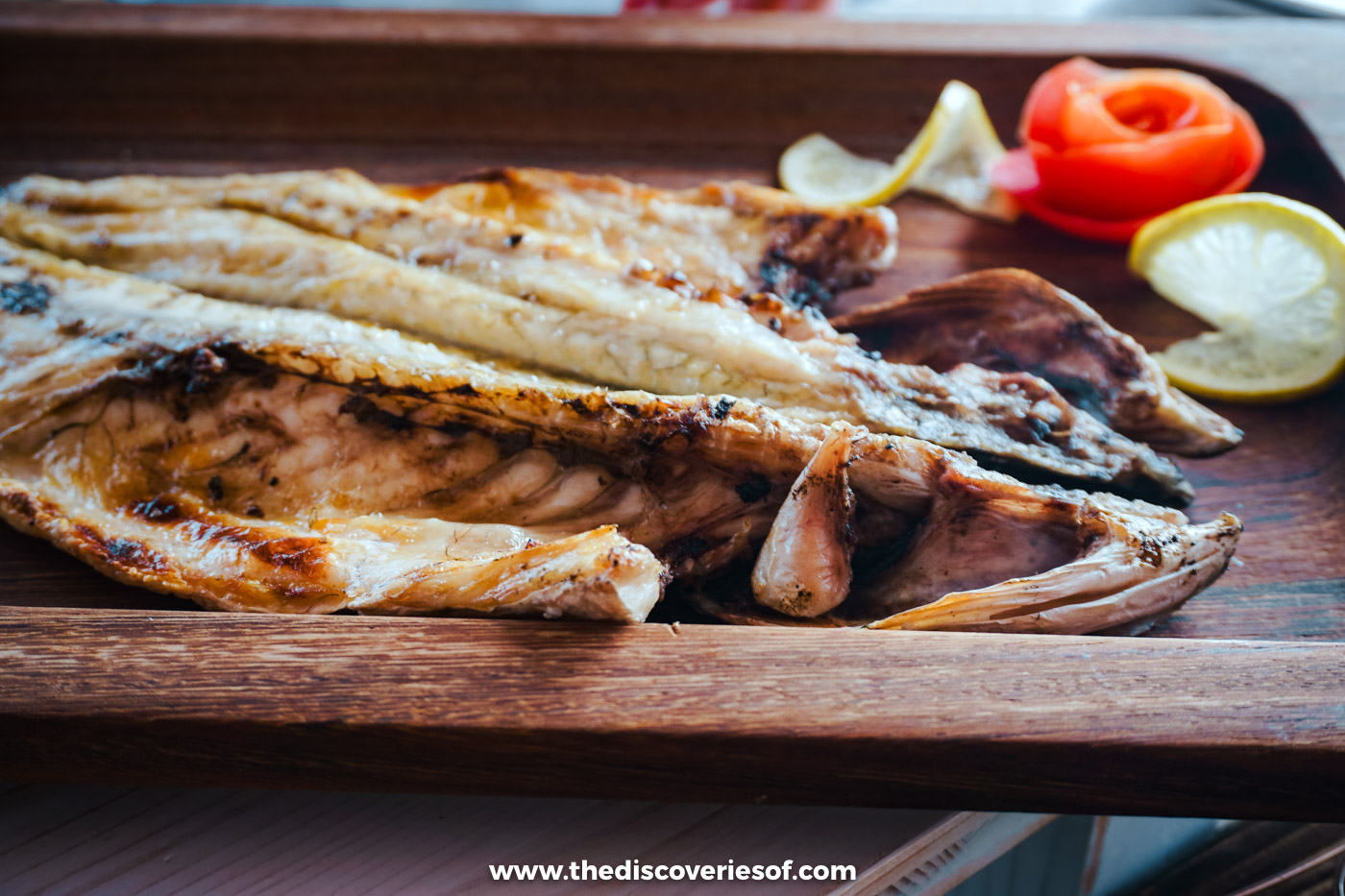 A whole grilled fish split open on a wooden board with lemon and tomato at Estaminé on Ilha Deserta