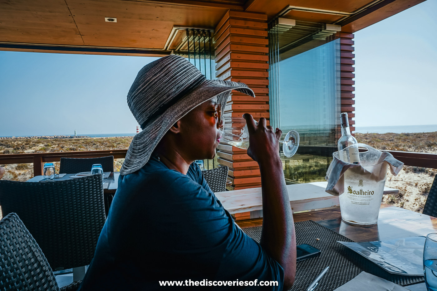 Julianna in a sun hat sipping white wine at a terrace table at Estaminé, with the Ilha Deserta dunes behind