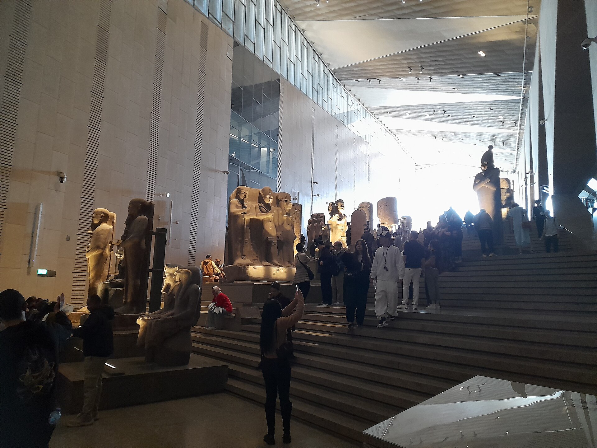 Visitors climbing the Grand Staircase of the Grand Egyptian Museum, lined with ancient statues under a glass wall
