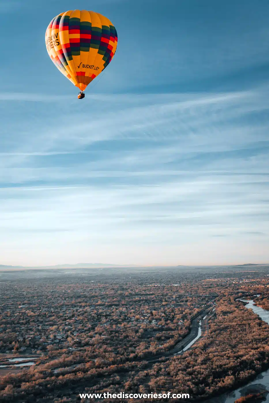 A red and yellow hot air balloon floating above the Rio Grande and its bosque at sunrise in Albuquerque