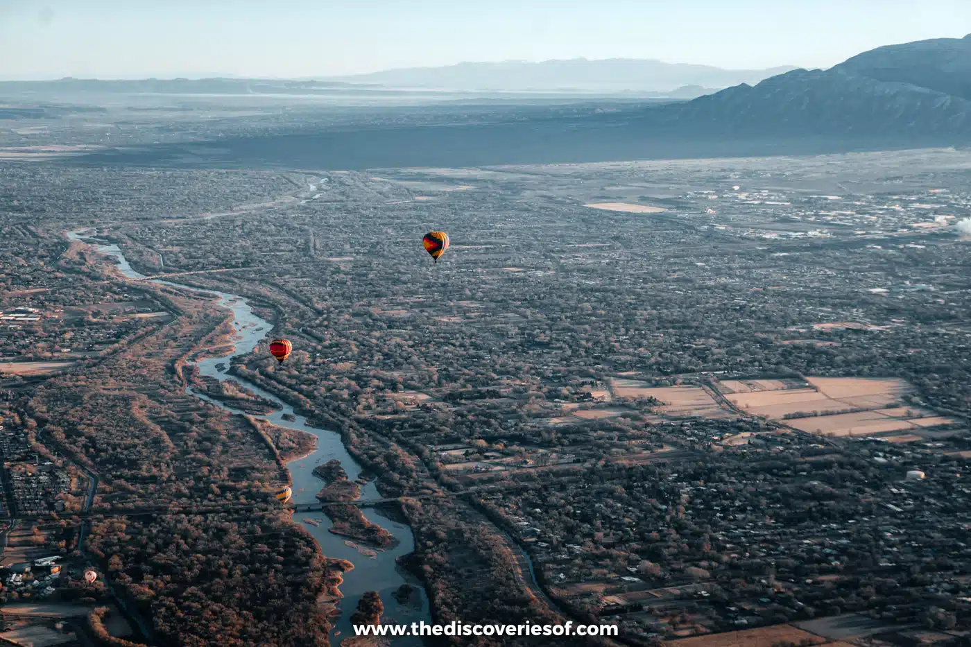 Hot air balloons drifting over the Rio Grande and its cottonwood bosque, with the Sandia Mountains behind Albuquerque