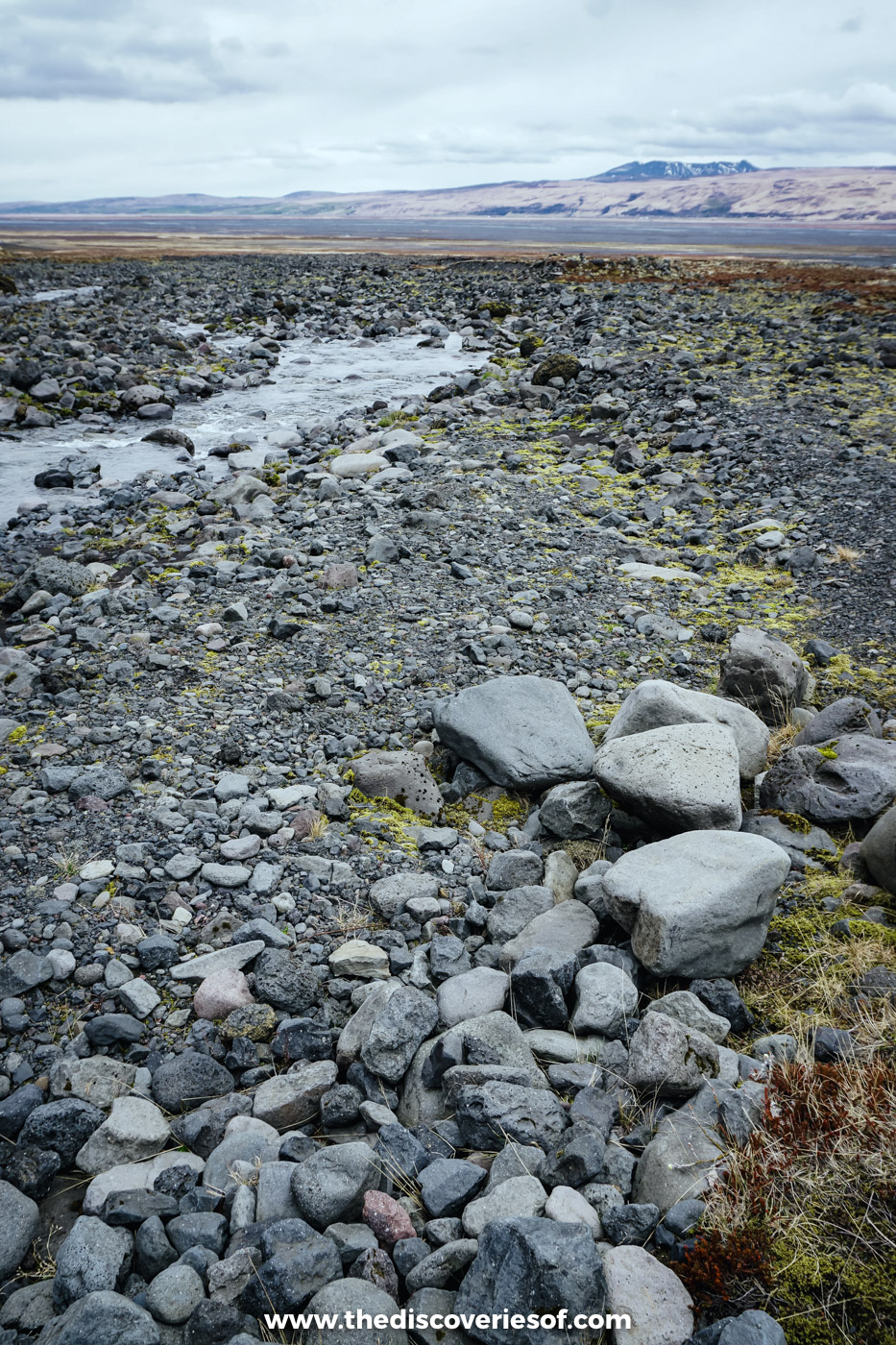 Meltwater running across a wide bed of grey stones and boulders below distant mountains in south Iceland