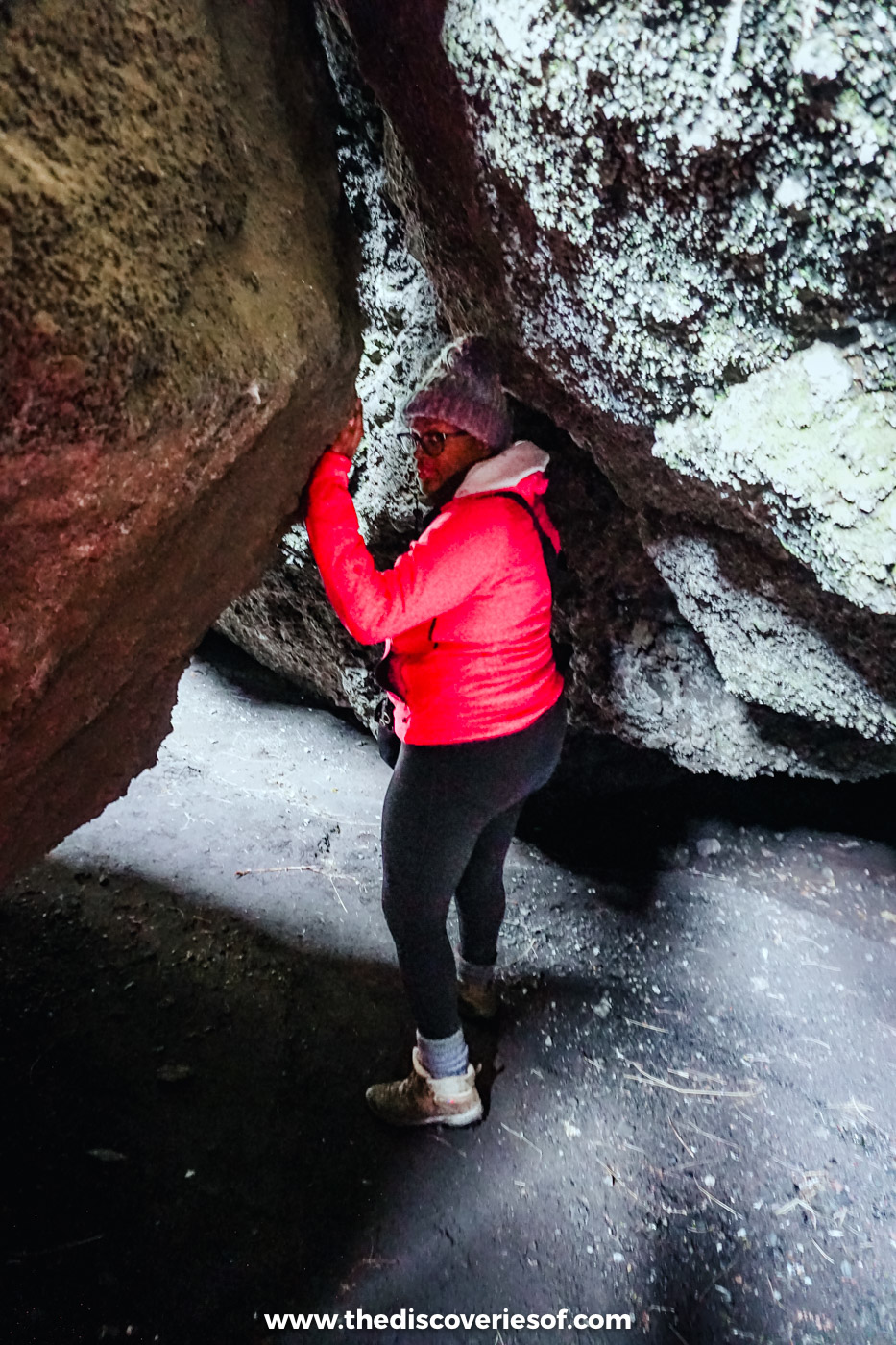 Woman in a pink jacket and bobble hat squeezing through a narrow gap between lichen-covered rocks near Þórsmörk