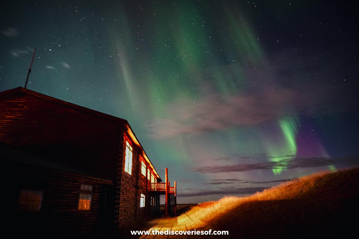 Green and purple Northern Lights over the lit timber buildings of Hotel Rangá near Hella at night