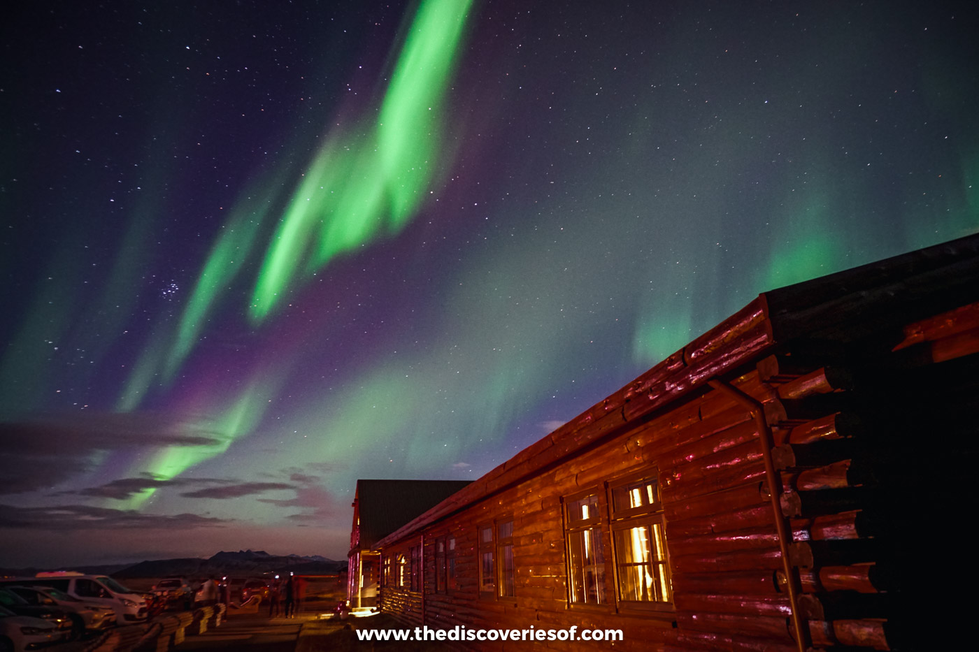 Green and purple Northern Lights swirling in the night sky above the timber wing of Hotel Rangá near Hella