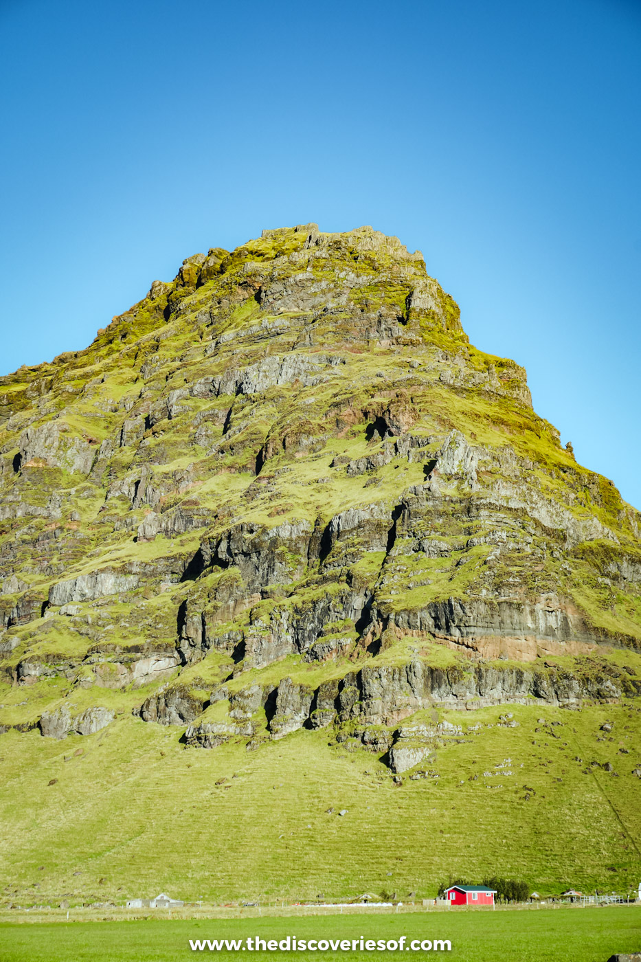 A moss-covered rock crag rising out of green fields under a blue sky on Iceland's south coast