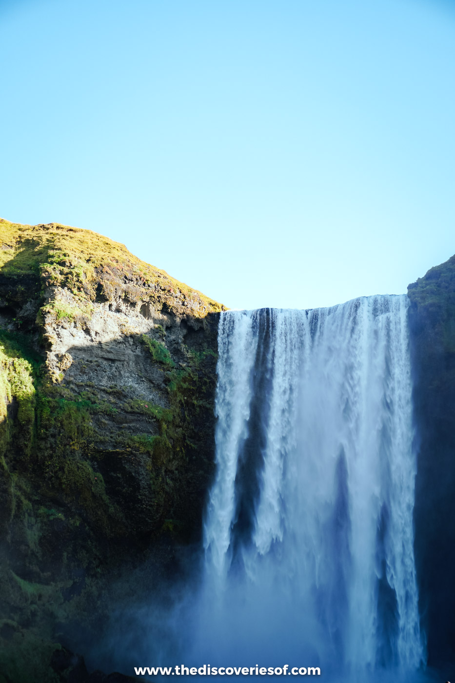Skógafoss pouring over a mossy green cliff under a cloudless blue sky in September