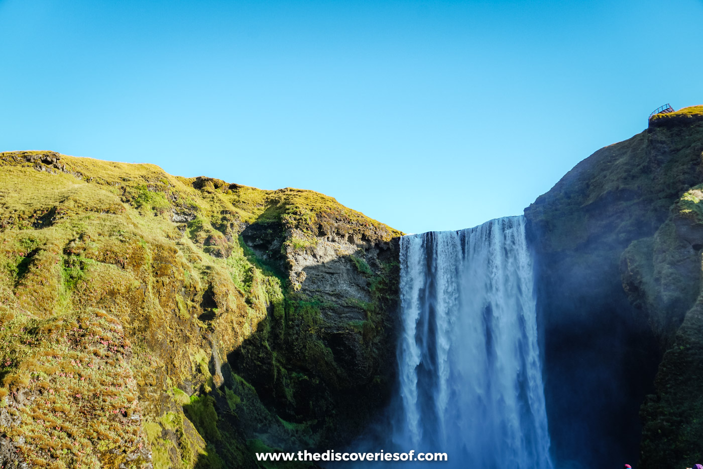 The broad curtain of Skógafoss between moss-covered cliffs, with spray drifting across the base